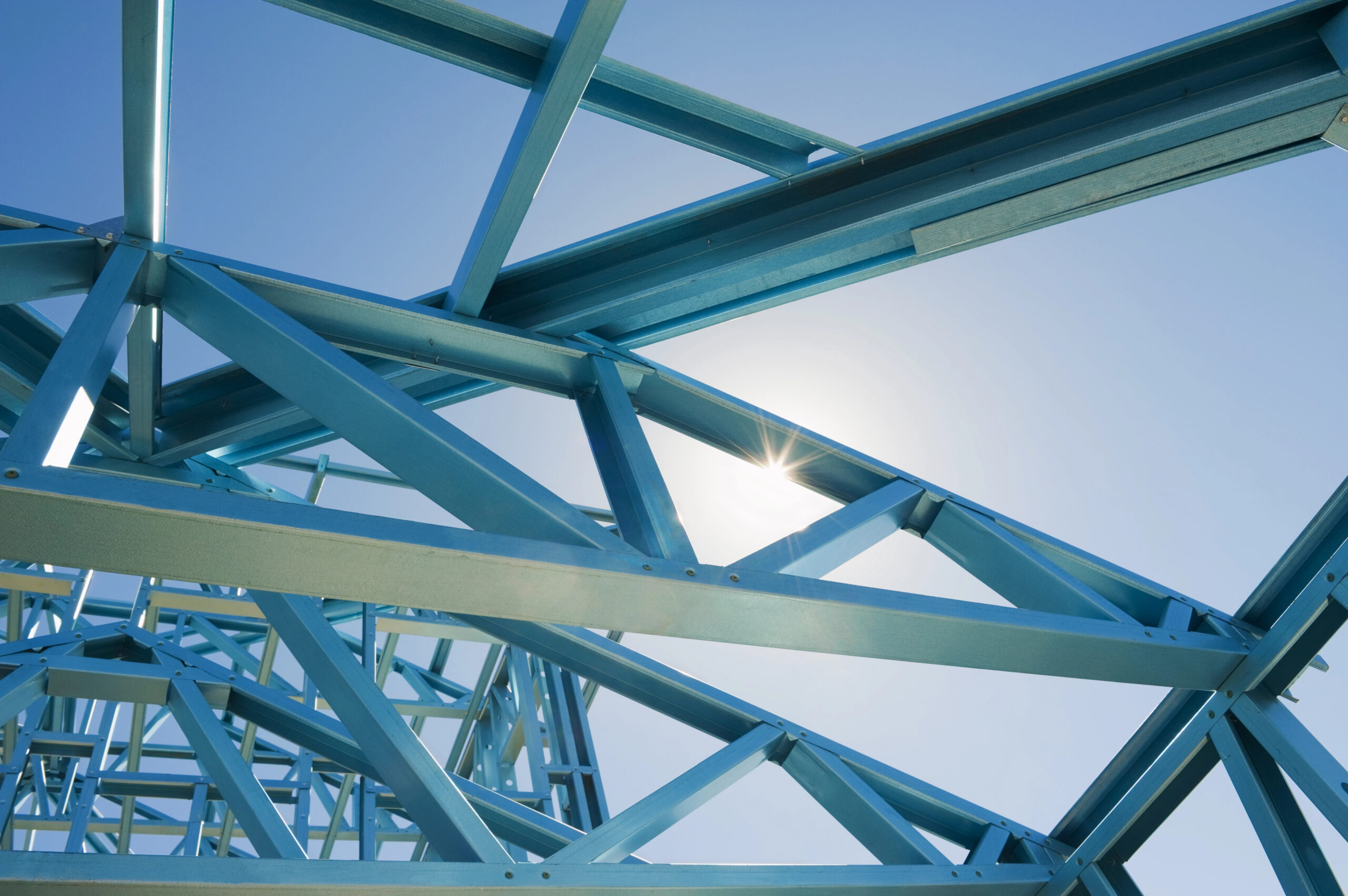 A skilled steel fabricator's craftsmanship is evident in the blue steel beams forming a complex geometric structure against the clear sky, with the sun peeking through gaps to create a bright starburst effect.