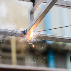 Close-up of a steel fabricator welding metal beams with intense sparks flying. A gloved hand steadies the tool, smoke rising from the heated area. The background is blurred, highlighting the precision craft involved in creating custom railings.