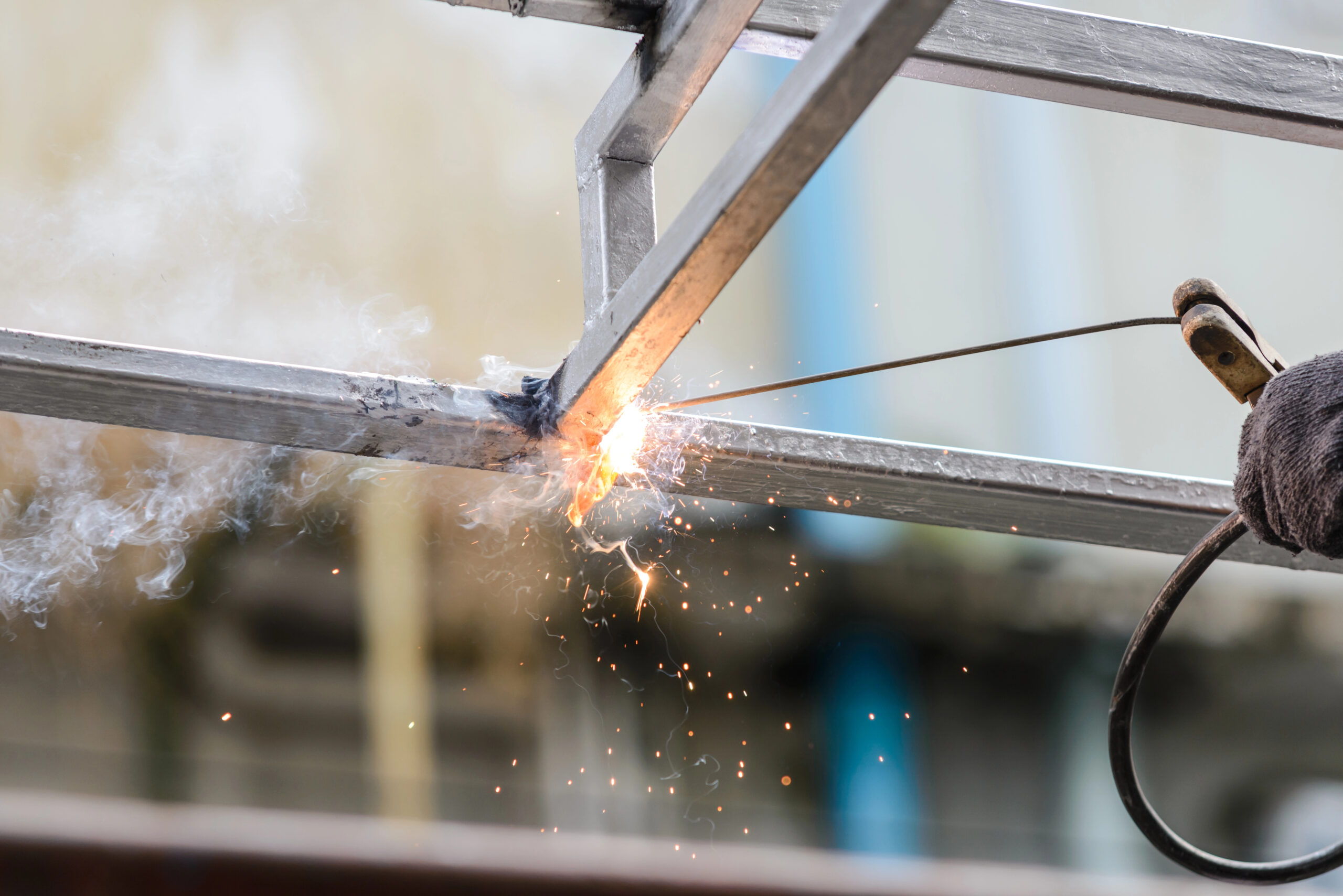 Close-up of a steel fabricator welding metal beams with intense sparks flying. A gloved hand steadies the tool, smoke rising from the heated area. The background is blurred, highlighting the precision craft involved in creating custom railings.