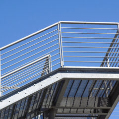 Metal staircase with custom railings and a grated platform, set against a clear blue sky. The design features horizontal bars and a sturdy steel frame, creating a modern industrial look reminiscent of sleek metal fire escapes.