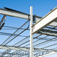 Steel beams and trusses form the skeletal structure of a building under construction, set against a clear blue sky. Nearby, custom railings are being crafted by skilled steel fabricators, indicating the early stages of architectural development.