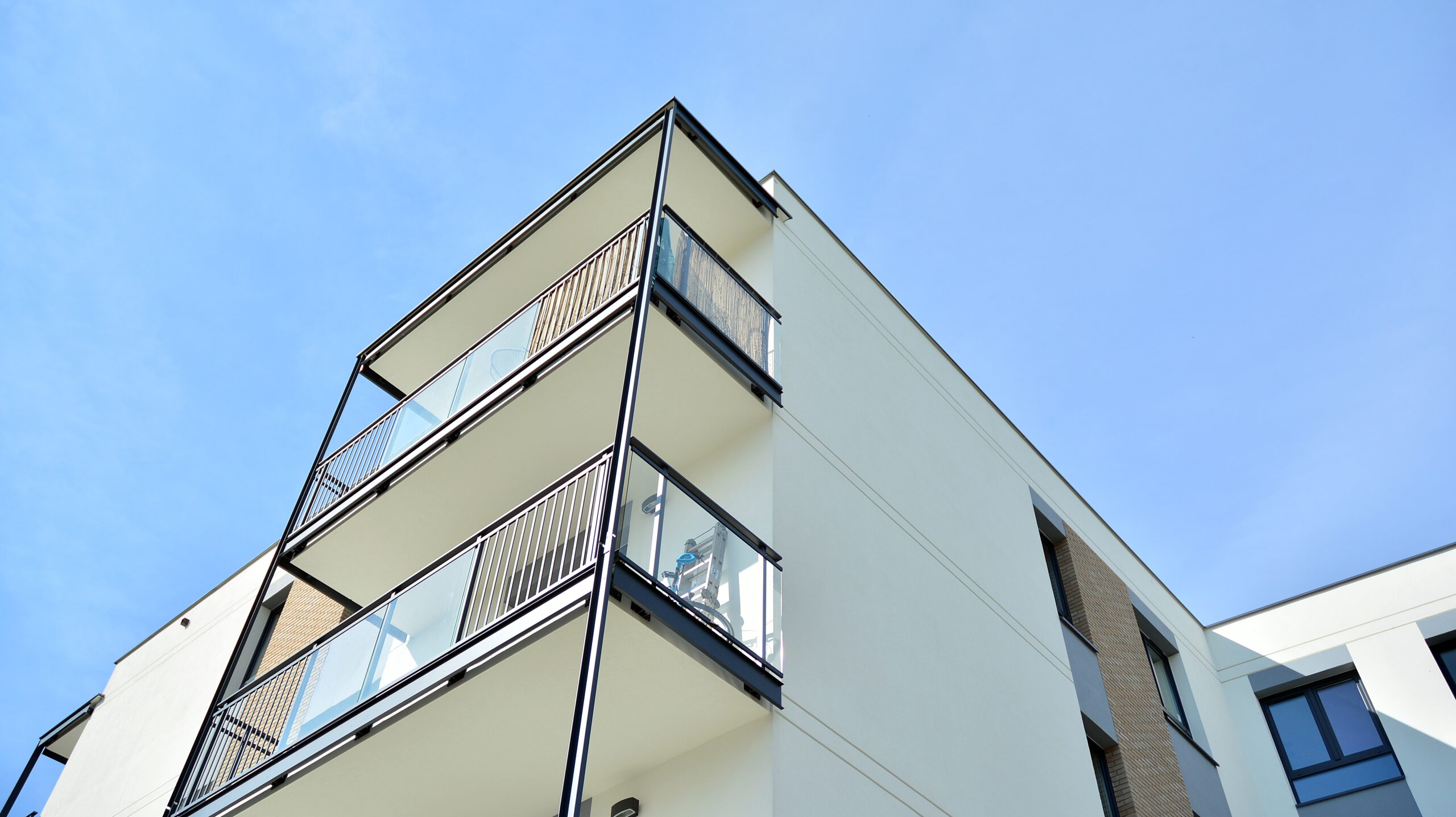 Modern apartment building with a white exterior and glass balconies under a clear blue sky. The photo captures the corner view, highlighting three floors with custom steel frame railings for a sleek, contemporary look.