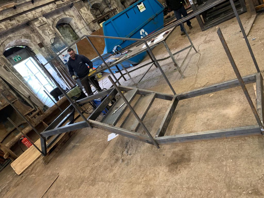 A steel fabricator stands in a bustling workshop, surrounded by metal frames on the floor. The industrial setting features custom railings, a blue recycling bin, wooden boards, and stone walls as the backdrop.