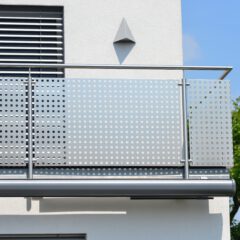 Modern balcony with custom railings made by a skilled steel fabricator, attached to a white building. A diamond-shaped wall light is above the railing, and there’s a window with horizontal blinds on the left and green tree branches on the right.