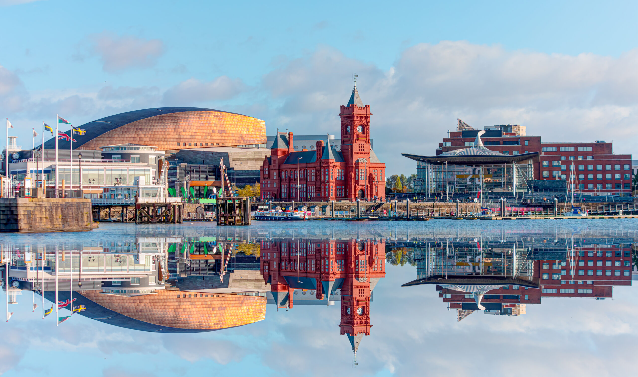 The image showcases a waterfront cityscape, featuring a prominent red brick building adorned with custom railings and a curved metallic structure. These architectural elements are beautifully reflected in the water, all under a clear blue sky with scattered clouds.
