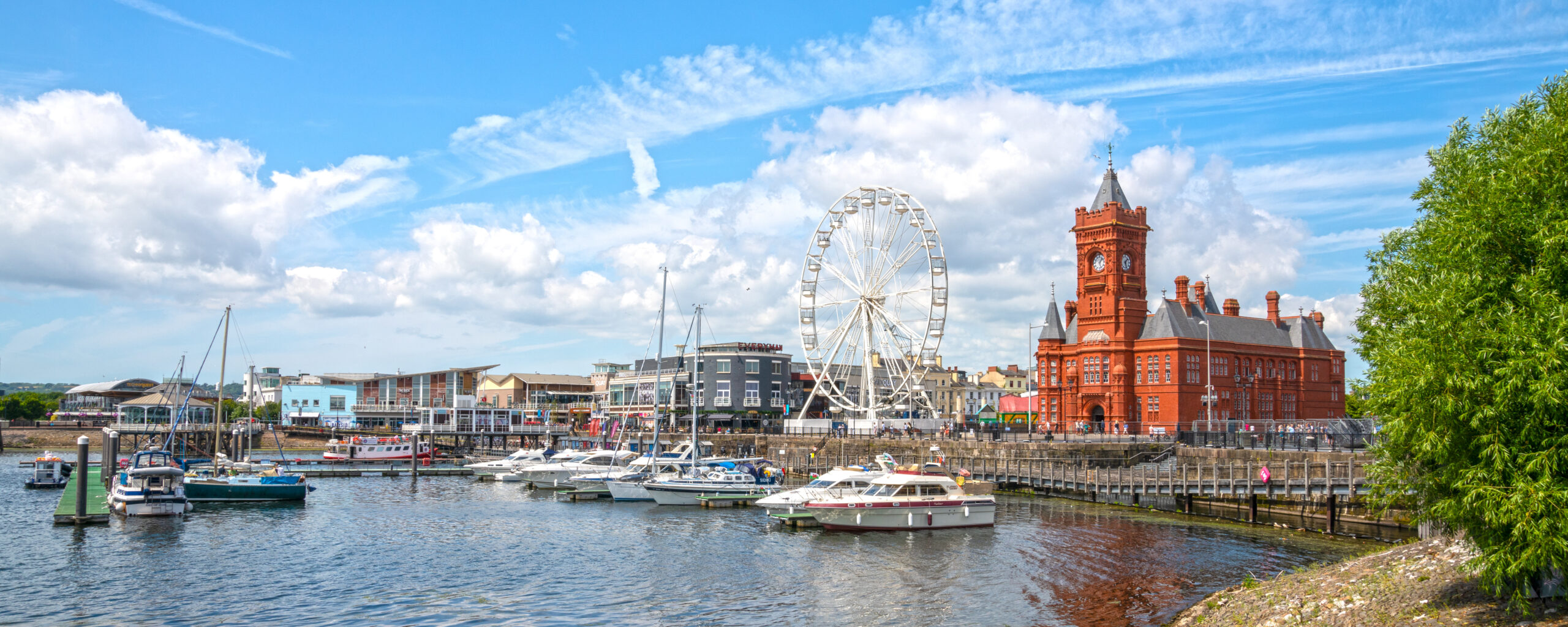 A waterfront scene with boats docked by the shore, a large Ferris wheel, and a distinctive red-brick building with a clock tower. Steel frame balconies blend seamlessly into the architecture, while scattered clouds fill the blue sky and trees frame the right side of the image.