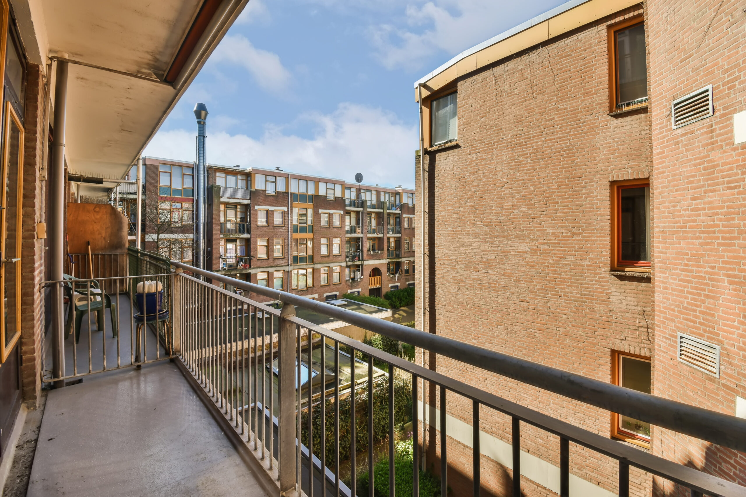 From a steel-frame balcony with custom railings, enjoy the view overlooking a courtyard embraced by brick apartment buildings. The metal railing encloses a cozy space with a small table and chairs. Sunlight filters through partly cloudy skies, bathing the scene in warmth.