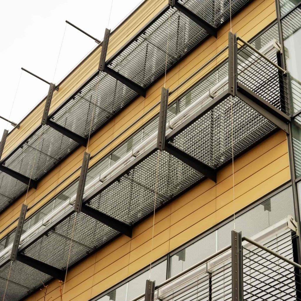 Close-up of a modern building facade featuring yellow horizontal siding and several metal mesh balconies, each supported by black frames and thin vertical cables.