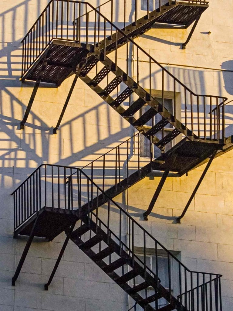 Black metal fire escape stairs zigzag up the side of a light-colored building, casting distinct shadows on the wall in warm sunlight.