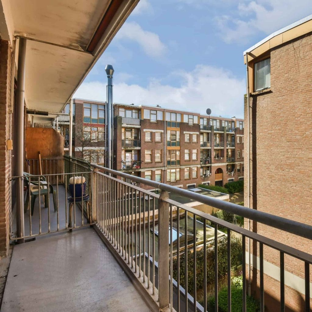 Residential balcony with metal railings overlooking a courtyard and neighboring apartment buildings under a blue sky with clouds.