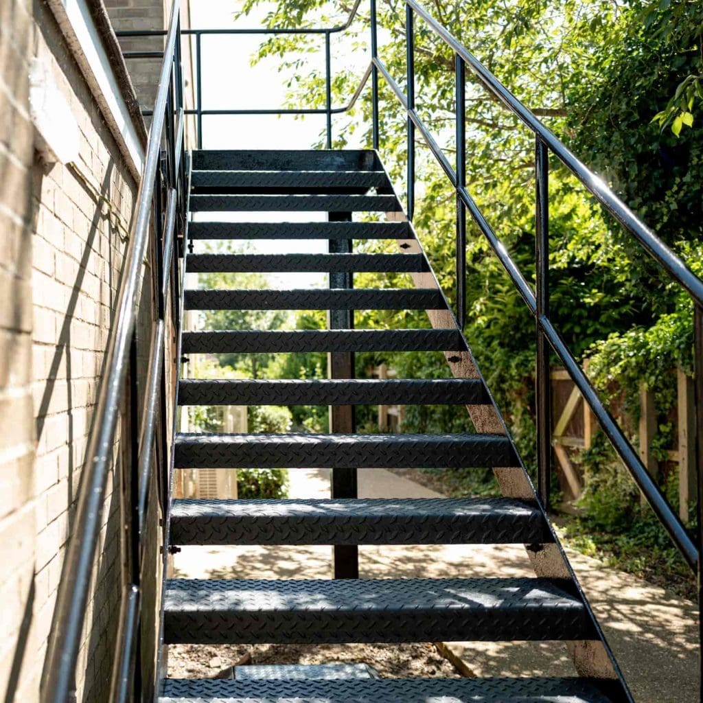 Metal outdoor staircase with black railings, attached to a brick building, leading upward with sunlight casting shadows, and green trees and a sidewalk visible to the right.