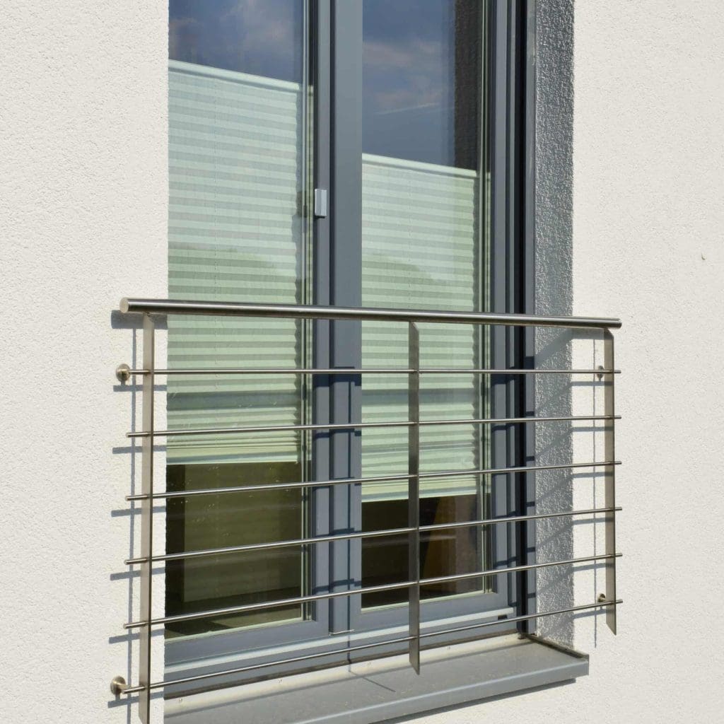 A modern glass window with closed blinds is shown. There is a simple metal railing installed outside the window, attached to a white exterior wall. Sunlight casts shadows on the wall and railing.